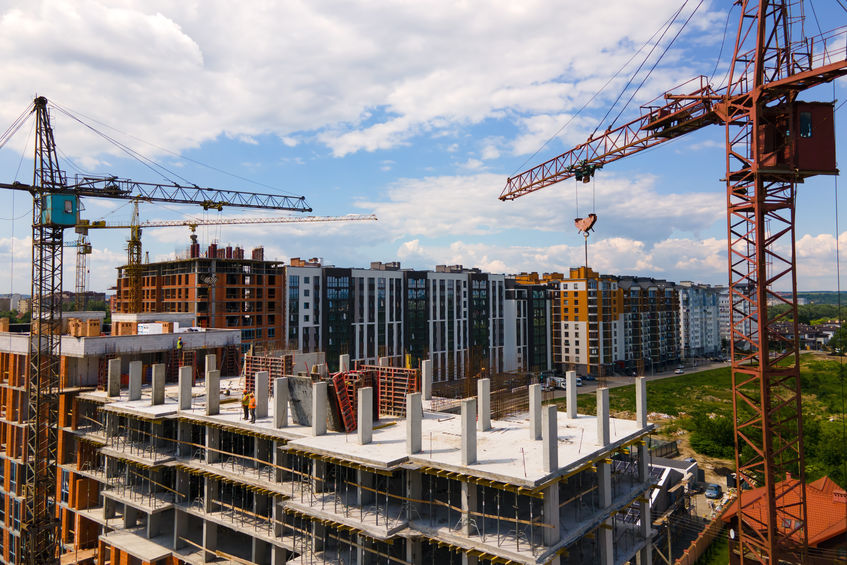Several cranes tower above a large construction site with multiple residential buildings under development under a blue sky with clouds.
