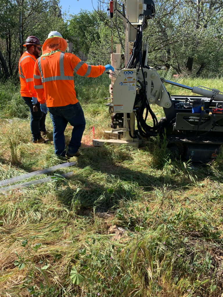 Two workers in orange safety vests operate a drilling rig in a grassy area surrounded by trees.