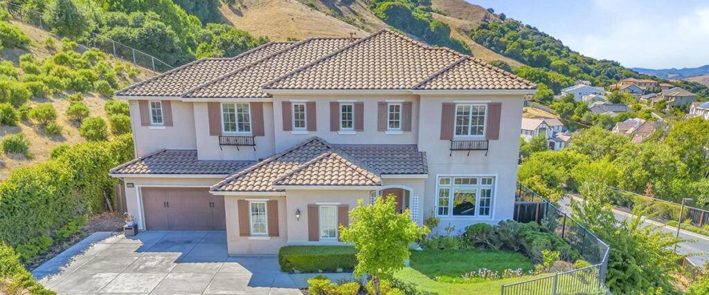 A two-story, tan stucco house with a tile roof, two-car garage, and landscaped front yard, set against a backdrop of green hills.