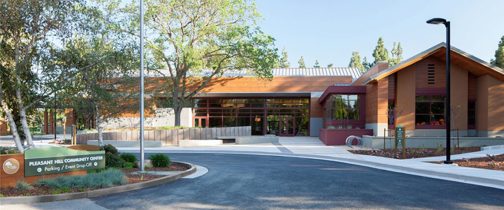 Entrance of Pleasant Hill Community Center with modern architecture, large windows, and a surrounding landscape of trees; a sign indicating parking and event drop-off is visible at the front.