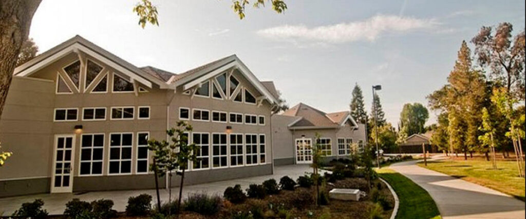 A modern building with large glass windows and a peaked roof, surrounded by a pathway and shrubbery, under a partly cloudy sky.