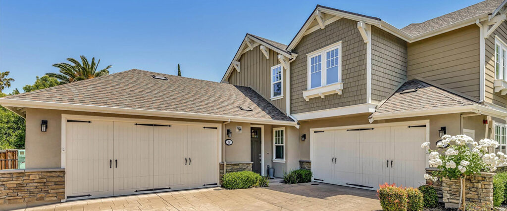 A two-story house with beige siding, white trim, and a gray shingled roof. The house has three garage doors and a paved driveway, with white flowers and greenery visible around the exterior.