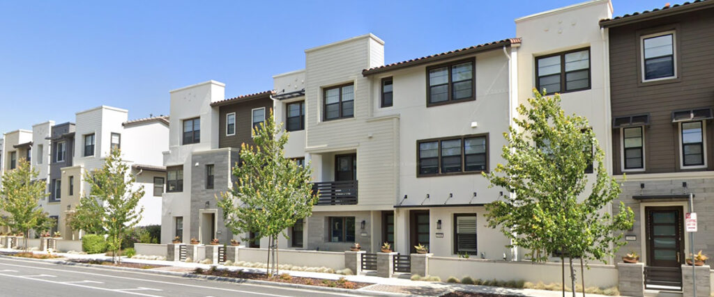 A row of modern three-story townhouses with balconies and trees lining the street under a clear blue sky.