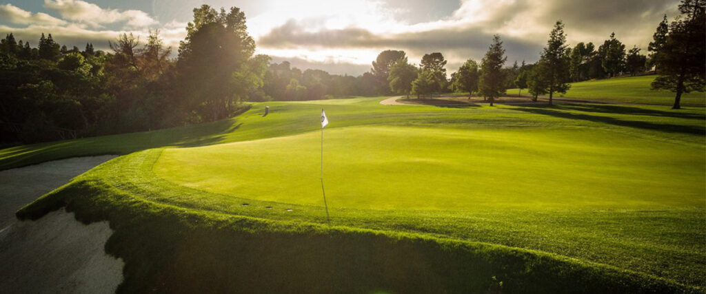 A well-maintained golf course with lush green grass, a sand bunker on the left, and a white flag marking a hole. Trees line the background under a partly cloudy sky.