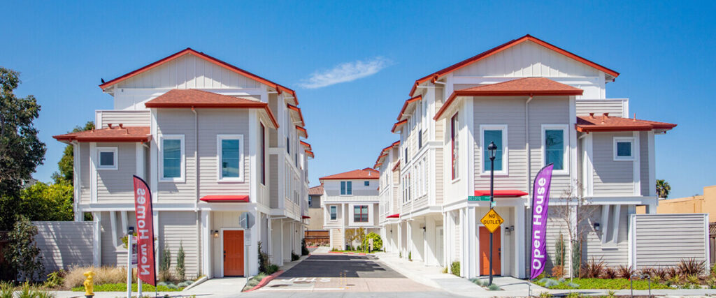 Two rows of white, three-story townhouses with red roofs stand under a clear blue sky. "Open House" signs are visible at the entrance of the complex, with a paved driveway between the buildings.