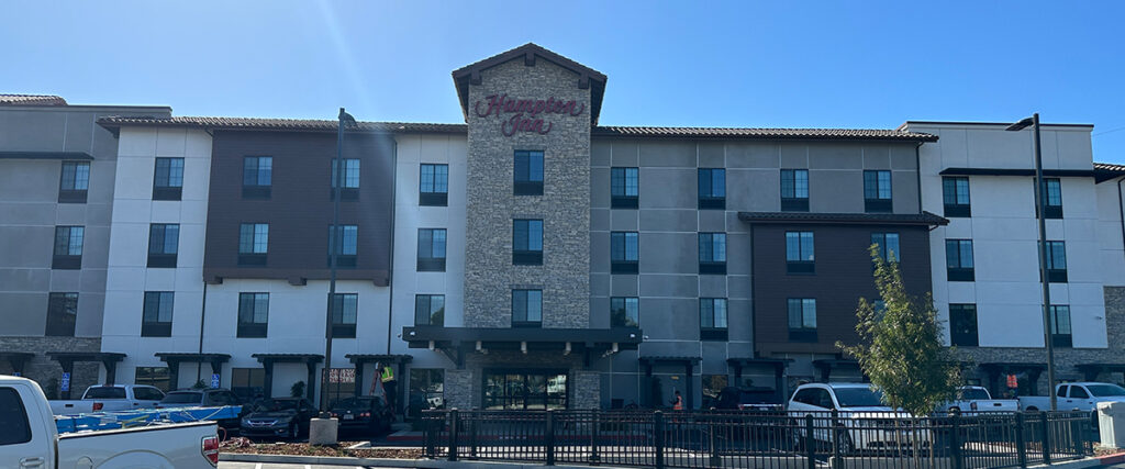 A multi-story Hampton Inn hotel building with a central tower entrance and several cars parked in front. The sky is clear and blue.