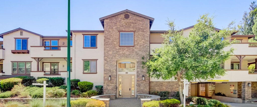 A three-story residential building with a stone facade, archway entrance, and balconies is surrounded by landscaped greenery and has a tree in the front yard. There's also a garage entrance on the right.