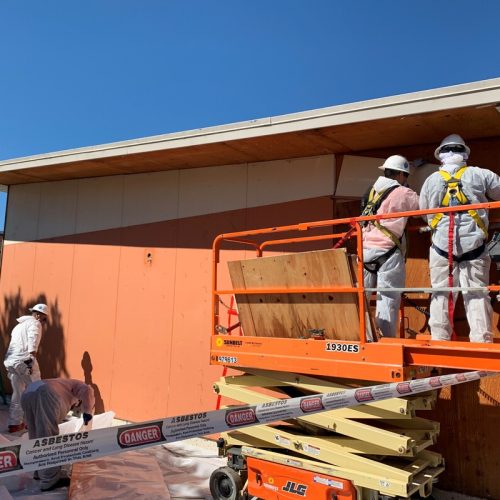 Workers in protective suits and helmets stand on a lift and work on an orange building. The area is marked with "Danger Asbestos" tape, indicating asbestos hazard precautions.
