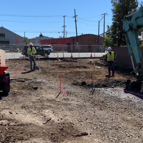 Construction workers operating heavy machinery and surveying at a construction site with exposed dirt and gravel. Cones and markers are positioned around the area.