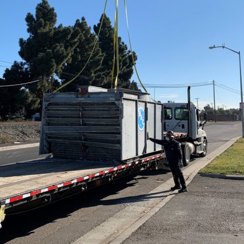 A large piece of equipment is being lifted onto a flatbed truck by a crane, while two workers supervise the loading process on a clear, sunny day.