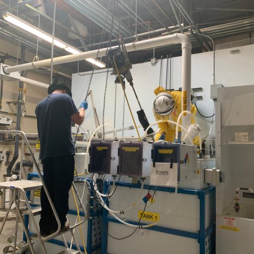 A person stands on a ladder, working with machinery and tanks in an industrial setting. Safety equipment and warning signs are present.