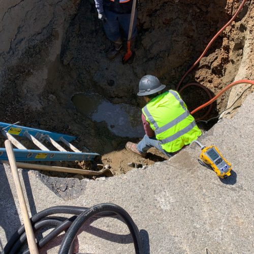 A construction worker in a neon yellow safety vest and helmet is working inside a large hole with equipment and ladders on the ground around them.