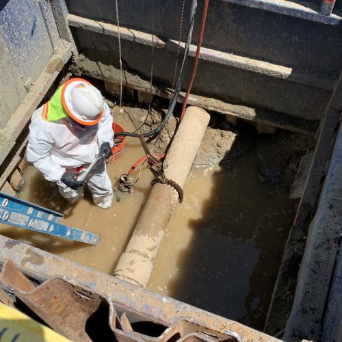 A worker in protective gear stands in a muddy trench, inspecting a large pipe connected to wires and equipment. A ladder is positioned on one side of the trench for access.