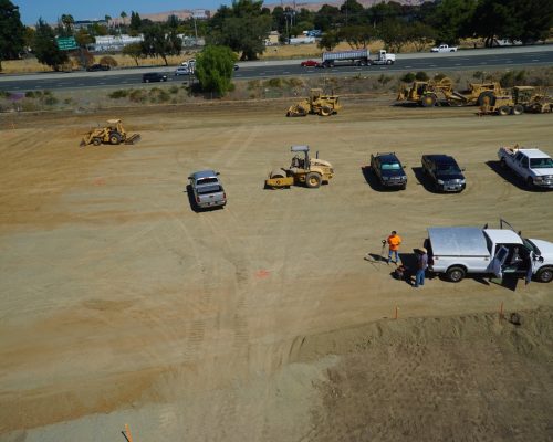 Construction site with vehicles and equipment on a leveled dirt area. Workers are near a white truck, with a highway and more vehicles in the background. Trees and hills are visible in the distance.