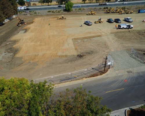 Aerial view of a construction site with several vehicles and construction equipment on a leveled dirt area, adjacent to a road and bordered by a fence and trees.