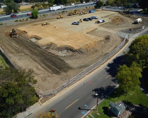 Aerial view of a construction site with a cleared and leveled dirt area, excavating equipment, a few parked vehicles, and a road running alongside the site.