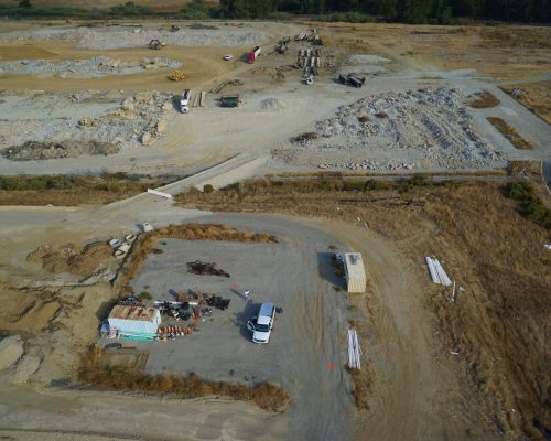 Aerial view of a construction site with earthmoving equipment, piles of materials, a white truck, and a work area with containers and supplies. Sparse vegetation covers the surrounding area.