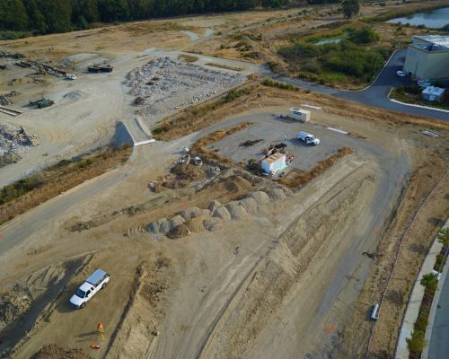Aerial view of a construction site with various machinery, dirt piles, and a few trucks. The area is surrounded by trees and includes a partially built structure.