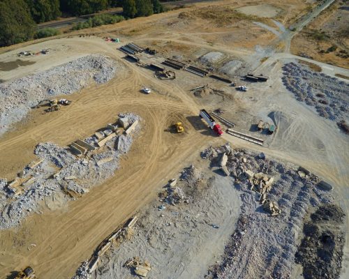 Aerial view of a construction site with heavy machinery, piles of rubble, and various construction materials scattered across a large, open area.