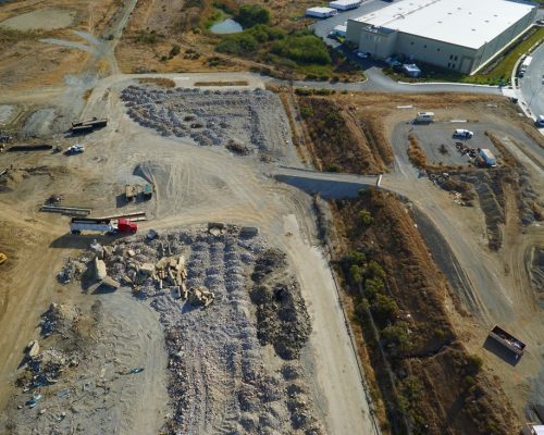 Aerial view of a construction site with materials piled on the ground, trucks and machinery visible, and a large warehouse building in the background.