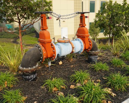 A large industrial valve system with red and blue components is installed outdoors amidst greenery and next to a building.