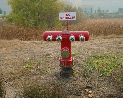A red fire department connection (FDC) sprinkler system stands in a grassy area, with a sign reading "FDC SPRINKLER." Industrial buildings are visible in the background.