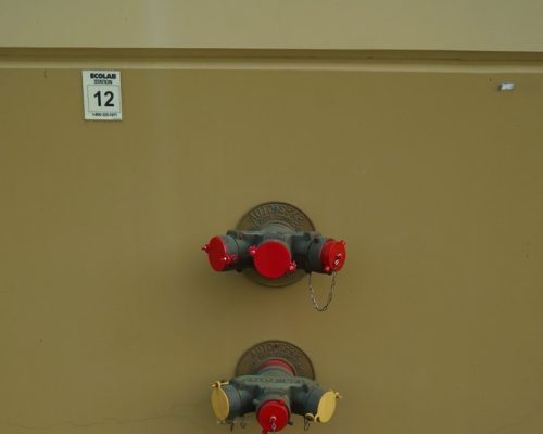 Two wall-mounted fire hose connections with red and yellow caps are attached to a beige external wall, under a small white sign labeled "Ecolab 12.